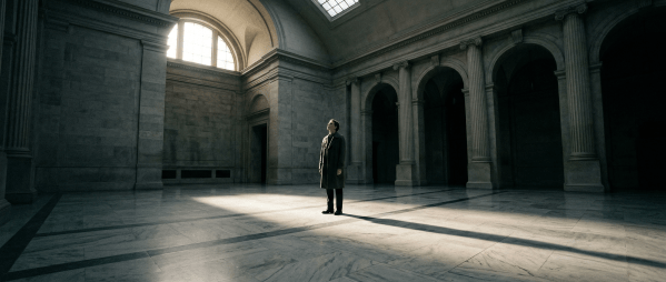 A solitary figure standing in a vast architectural space at dusk, light casting long shadows across the floor, looking forward with quiet determination.