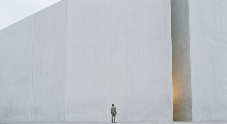 A tiny figure seen from behind at the foot of an enormous smooth pale wall that extends beyond the frame, looking up.